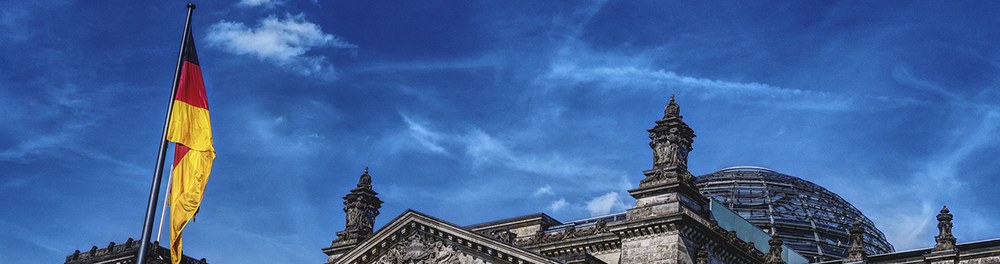 Slider: Kuppel des Bundestages vor blauem Himmel mit Deutschlandfahne im Vordergrund