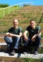 Andreas Gattinger und Christian Lambertz sitzen auf einer grasbewachsenen Treppe und schauen in die Kamera