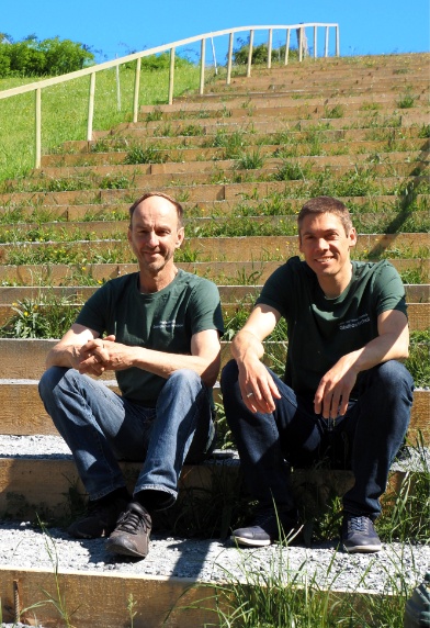 Andreas Gattinger und Christian Lambertz sitzen auf einer grasbewachsenen Treppe und schauen in die Kamera