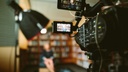 Sie sehen eine Fotografie zum Thema Videocast. Zu sehen ist eine Videokamera, die eine Person fokussiert.
