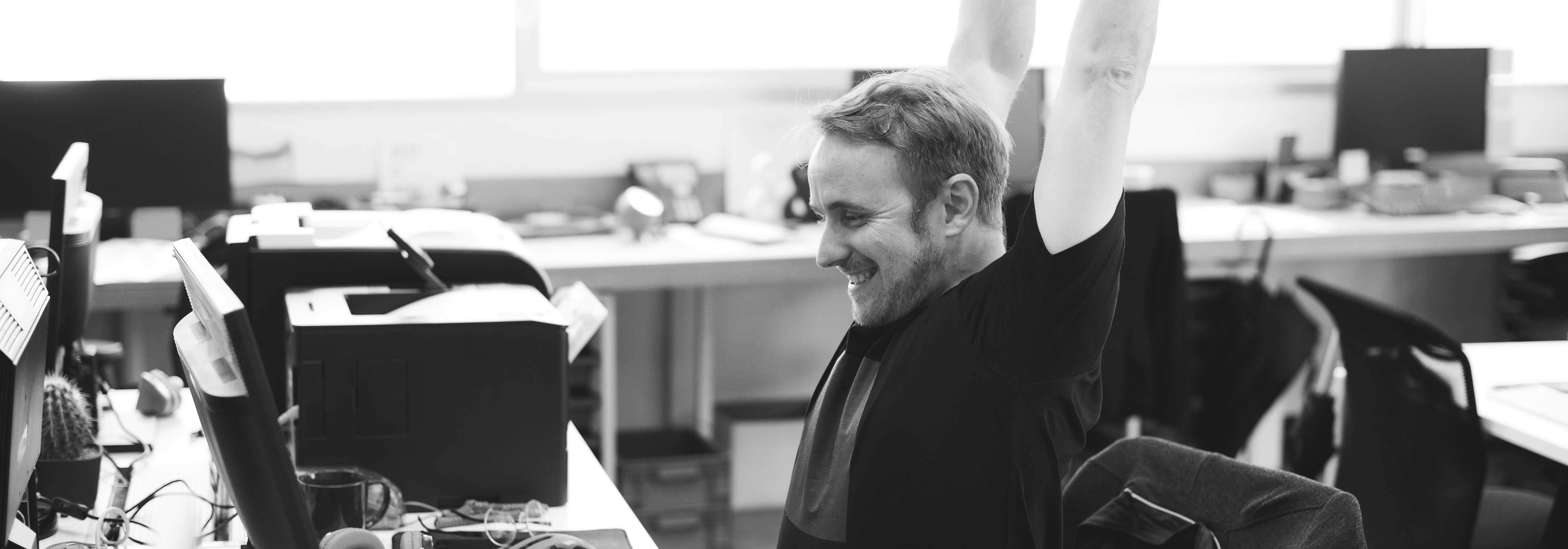 Slider: lächelnde Person an Schreibtisch in einem Büro, die sich streckt (Bildbeschreibung und Bildnachweis siehe Quellenverzeichnis) / Slider: smiling person stretching in front of a desk in an office setting