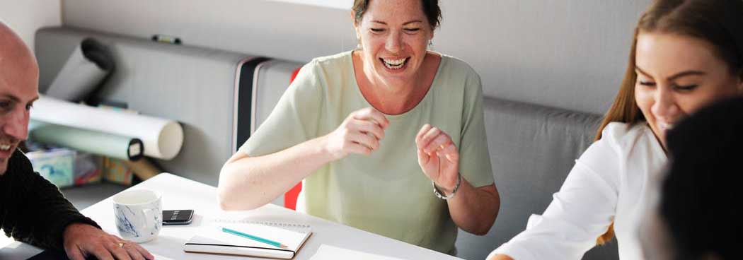 Slider: lachende Frau an Tisch mit Gruppe von Menschen, die sich unterhalten (Bildbeschreibung und Bildnachweis siehe Quellenverzeichnis) / Slider: laughing woman on a table, talking with a group of people