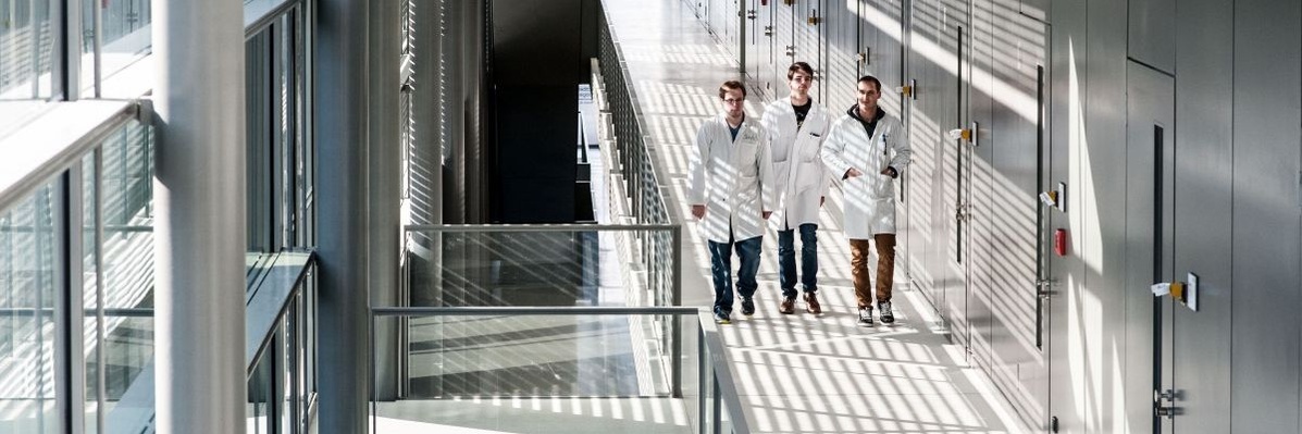 Schmuckgrafik: Drei Personen in weißen Laborkitteln laufen in einem Laborgebäude über einen Gang 