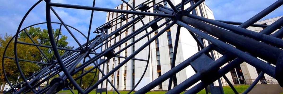 Schmuckgrafik: Eine große Metallkonstruktion aus Röhren, Ringen und Verstrebungen auf eine Wiese vor dem Institutsgebäude.