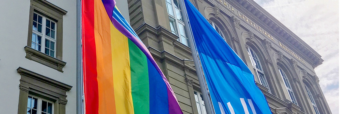 Symbolfoto: Am Fahnenmast vor dem Uni-Hauptgebäude wehen eine Regebogenflagge und die blaue Flagge der JLU.