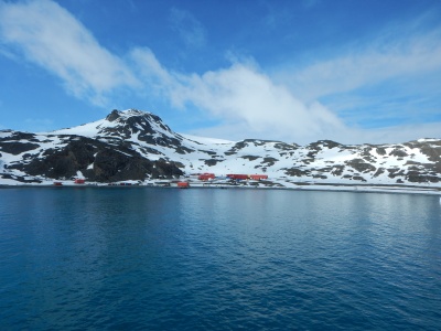 Eine Forschungsstation mit roten Gebäuden in der Antarktis, umgeben von schneebedeckten Bergen unter blauem Himmel.