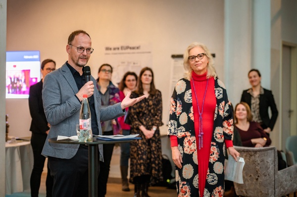 EUPeace_Rede.jpg Ein Mann im Anzug mit Mikrofon in der Hand, rechts von ihm steht eine Frau, im Hintergrund weitere Zuhörende.
