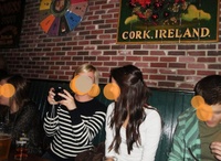 Versuchtsbild Vier Personen sitzen in einem Pub an einer Ziegelwand. An der Wand hängen dekorative Schilder, darunter eines mit der Aufschrift „CORK, IRELAND“. Die orangefarbenen Kreise auf den Gesichtern symbolisieren die Blickrichtungen der Testpersonen.