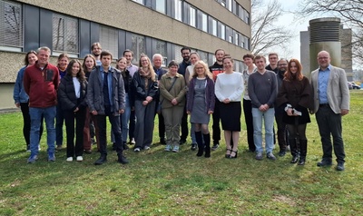 Auf einer Wiese stehen vor einem mehrstöckigen Universitäts-Gebäude 23 Frauen und Männer und lächeln in die Kamera. 