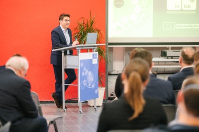 Ein Mann steht an einem Rednerpult und spricht. Im Vordergrund ist das Publikum von hinten zu sehen.