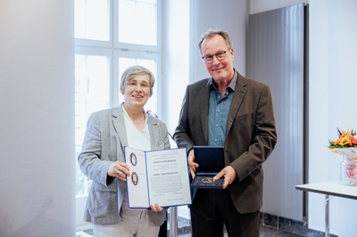 Eine Frau präsentiert die offizielle Urkunde, während ein Mann die dazugehörige Medaille in einer blauen Box hält.