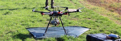 Eine große, professionelle Multicopter-Drohne steht auf einem schwarzen Untergrund auf einer grünen Wiese. Im Hintergrund sind die Beine einer Person zu sehen.