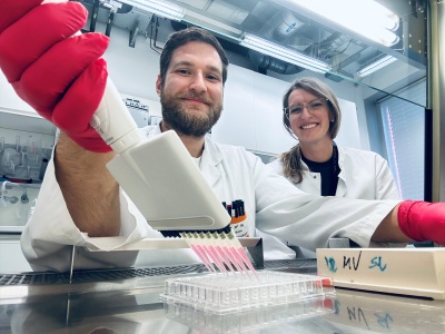 Zwei Forschende in weißen Kitteln sind im Labor. Einer füllt mit einer Mehrkanal-Pipette eine rosa Flüssigkeit in kleine Behälter.