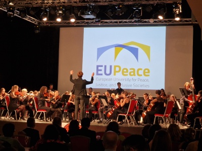 Auf einer Bühne spielt ein studentisches Orchester. Der Dirigent hat beide Arme erhoben. Im Hintergrund ist das EU Peace Logo zu sehen.
