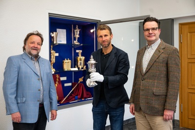 Drei Männer stehen vor dem geöffneten Vitrinenschrank. In der Mitte hält Prof. Krüger den Pokal mit Handschuhen.