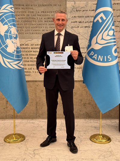 Der Professor hält die Urkunde mit beiden Händen vor sich. Hinter ihm sind zwei Fahnen der Vereinten Nation und der FAO zu sehen. Er hat einen schwarzen Anzug an.