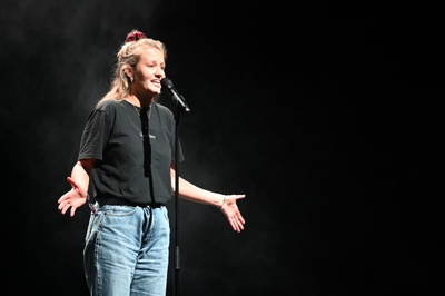 Leah Weigand steht vor einem schwarzen Hintergrund, spricht in ein Mikrofon und hat beide Hände leicht erhoben.