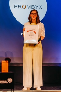 Fabiola Neitzel steht auf der Bühne und hält ihre Urkunde strahlend vor sich. Im Hintergrund ist das Logo von Prombyx zu sehen.