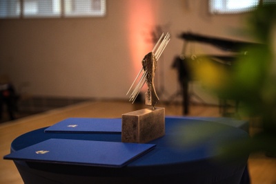 Der Preis steht auf einem Tisch. Daneben liegen blaue Urkundenmappen. Im Hintergrund sieht man den Klavierflügel der Universitätsaula.