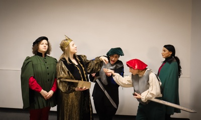 Fünf Schauspielerinnen als Männer verkleidet in einer Szene: In der Mitte ein König, der Geld an zwei Ritter mit langen Schwertern verteilt. Rechts und links stehen zwei Begleiter und schauen ernst zu.
