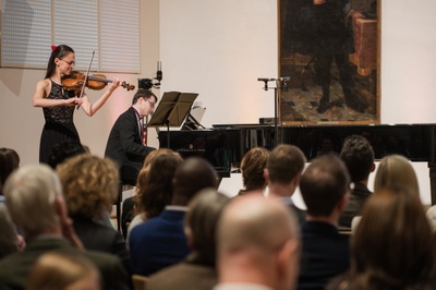Eine Frau spielt Violine. Am Flügel sitzt ein Pianospieler. Im Vordergrund Publikum von hinten.