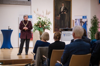Frau Lorenz steht auf der Bühne. Sie trägt die Amtskette. Vor ihr sitzt Publikum. Im Hintergrund ist ein Blumenbukett.