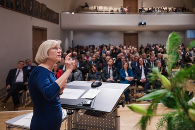 Blick von der Bühne ins Publikum. Im Vordergrund steht die Festrednerin. Sie dreht sich zur Kamera und schaut auf Ihre Folienwand.