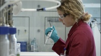 Die Forscherin arbeitet im Labor mit einer Pipette. Sie trägt einen roten Laborkittel, Schutzbrille und türkise Einweghandschuhe.