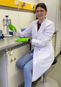 Die Forscherin trägt einen weißen Laborkittel und grüne Einweghandschuhe. Sie sitzt im Labor auf einem Hocker und hälte eine Pipette in der Hand.