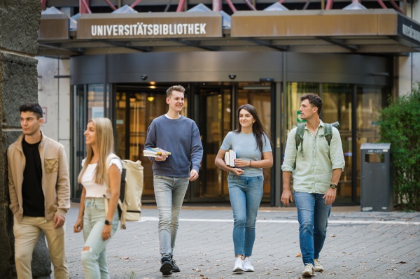 Fünf Studierende - 2 Frauen und 3 Männer - kommen aus der UB. Zwei Personen tragen Bücher - im Hintergrund ist die Drehtür der UB zu sehen.