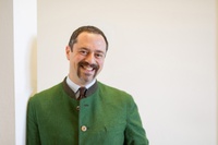 Porträtfoto Oberkörper: Mann mit braun-grau melierten Haaren und Bart lehnt an einer hellen Wand und blickt lächelnd in die Kamera. Er trägt ein weißes Hemd und eine braune Krawatte, darüber einen grünes Wollsakko mit Holzknöpfen.