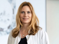 Porträtfoto: Frau mit langen blonden Haaren und silberner Kette blickt lächelnd in die Kamera. Sie trägt ein schwarzes Oberteil, darüber einen weißen geöffneten Kittel. Heller Hintergrund.