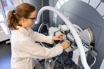 Junge Frau mit braunem Haar, das zu einem Zopf gebunden ist, steht an einem Großgerät und hält zwei Pole einer Batterie in den Händen. Die Frau trägt eine Schutzbrille und einen weißen Kittel.