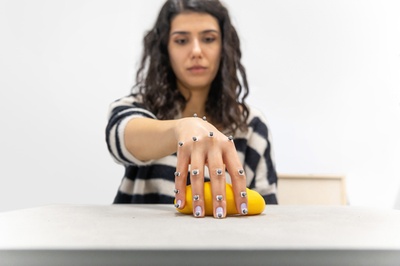 Junge Frau mit dunklen lockigen Haaren sitzt frontal gegenüber der Kamera an einem Tisch. Die Frau hält eine Plastikbanane in der Hand. An ihrer Hand sind mehrere Elektroden befestigt, der Blick der Frau ist auf die Banane gerichtet.
