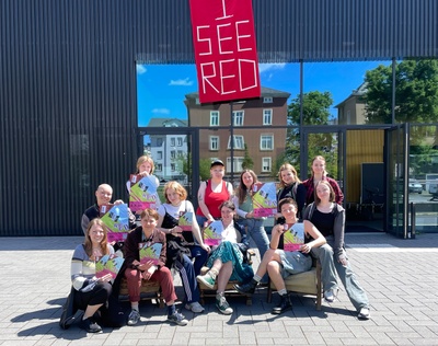 Zwölf Studierende auf und um Sessel auf dem Platz vor dem Theaterlabor in der Sonne. Einige halten das Plakat der Theatermaschine in den Händen. Am Theatelabor hängt ein rotes Banner mit der Aufschrift "I see red".