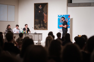 Ein Mann steht vor einer dreiköpfigen Jury und hält seinen Vortrag. Im Hintergrund das Plakat des Idea Slam 2024.