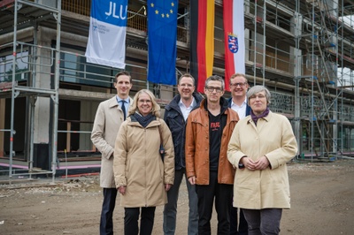 Sechs Personen stehen vor dem Rohbau des Seminargebäudes. Am Gebäude hängen die Flaggen der Uni, der EU, Deutschlands und Hessens.