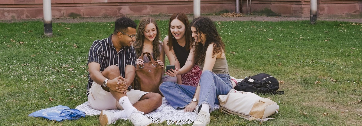 Schmuckbild: Vier Studierende sitzen auf einer Wiese und schauen sich gemeinsam einen Smartphone-Bildschirm an
