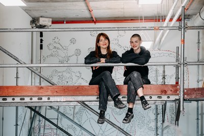 Studentinnen sitzen auf dem Gerüst vor der fertigen Wandbemalung und lächeln in die Kamera